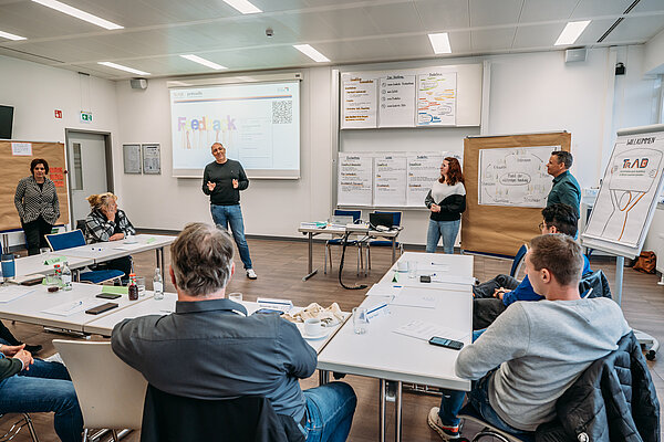 Begrüßung Teilnehmende der TrAB Inhouse-Schulung sitzen in einem Seminarraum in U-Form, während Provadis Geschäftsführer Holger Kison im Vordergrund steht und die Teilnehmenden begrüßt. Man sieht Inhalte zur pädagogischen Qualifizierung; Flipcharts und eine Präsentationsfolie verdeutlichen den praxisnahen Workshop-Charakter.