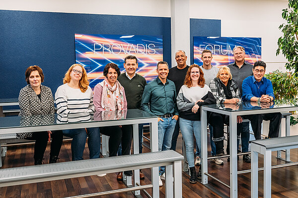 Teamfoto in einem modernen Büro mit buntem Hintergrund Gruppenfoto der Teilnehmenden und Projektbeteiligten der TrAB-Inhouse-Schulung bei Infraserv Höchst, die gemeinsam in einem modernen Raum stehen und lächeln, im Hintergrund sind Bildschirme mit der Aufschrift „Provadis Forum“ zu sehen.